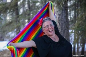 woman in  a black top, and a rainbow fabric stretched out behind her. She is standing in the woods.