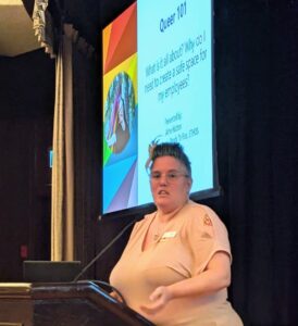 woman in a pink top standing at a podium speaking with a powerpoint slide behind her that says Queer 101